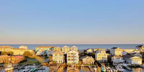 Sunlit coastal houses and docks with calm water and a visible moon in the sky.