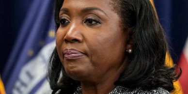A serious woman speaking at a press event with flags in the background.