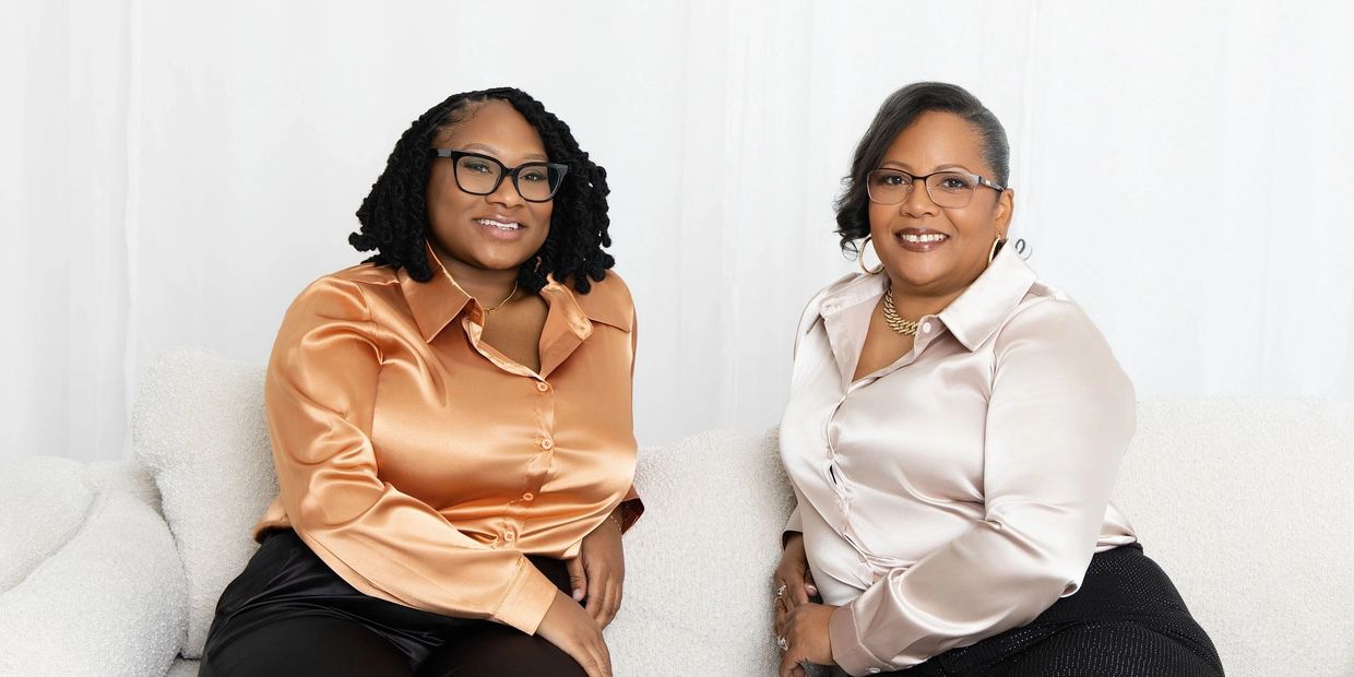 Two women in elegant satin blouses and glasses sit smiling together on a white couch.