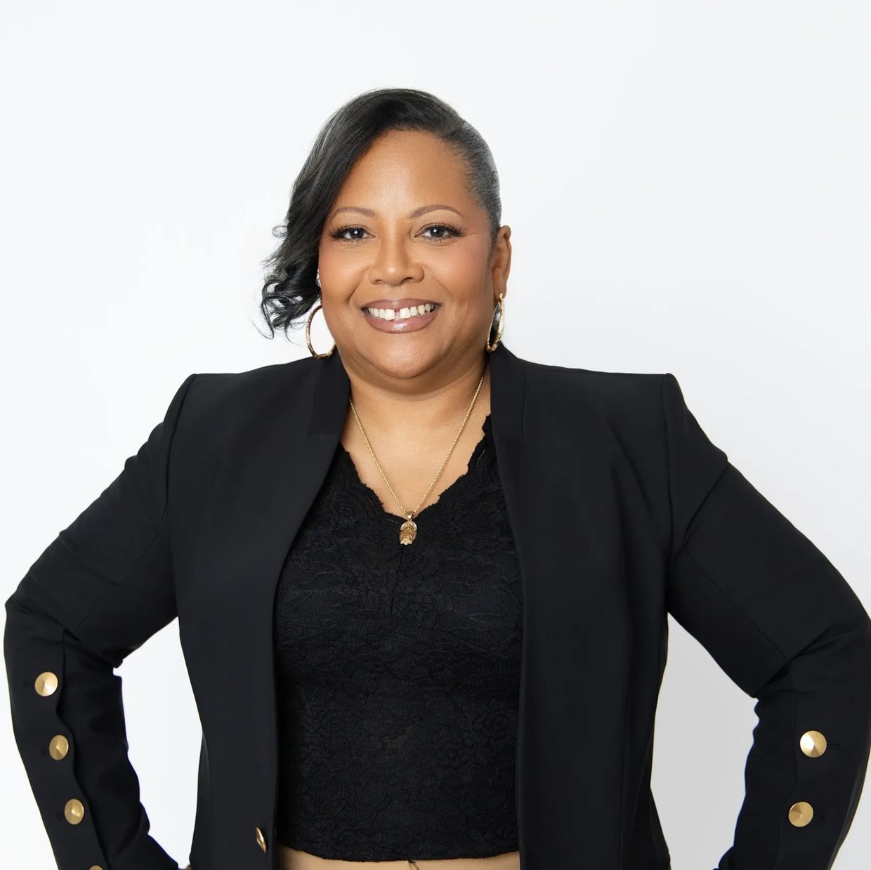 Confident woman smiling, dressed in black with gold accessories against a white background.