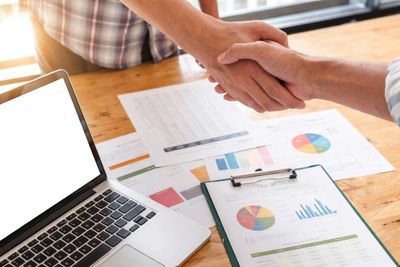 Two people shaking hands over business documents and a laptop.