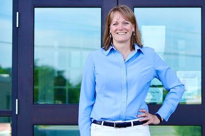 A smiling woman in a blue shirt stands confidently outside glass doors.