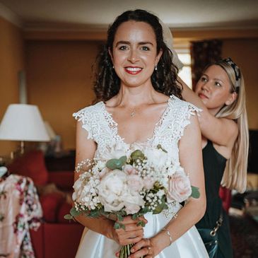 Bride holding bouquet with bridesmaid adjusting her dress behind.