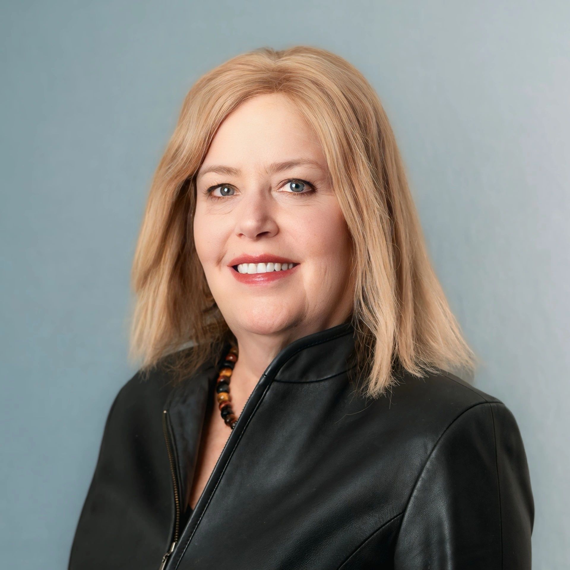 Smiling woman with blonde hair wearing a black leather jacket and beaded necklace.