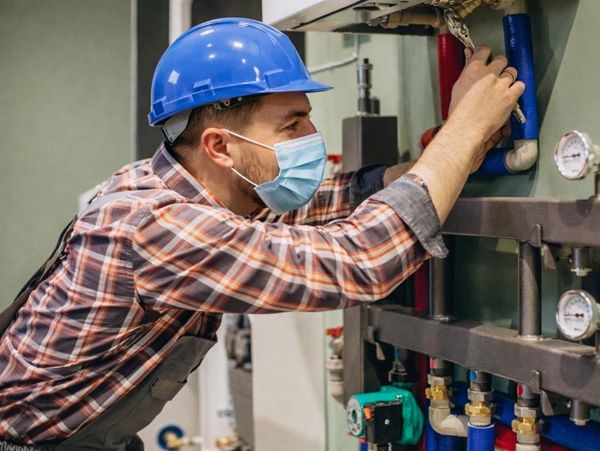 A technician in a blue hard hat and mask inspecting pipes for Furnace Repair in Stillwater.