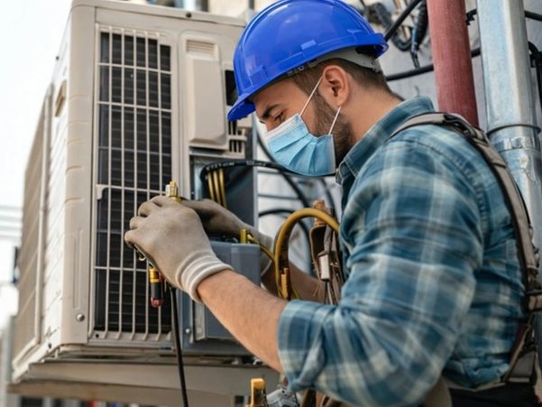 A technician in a blue hard hat and mask repairing an outdoor AC unit. Call AirZ Heating and Cooling