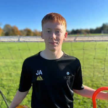 Young man in sports attire holding a yellow soccer ball and orange training equipment on a sunny day.
