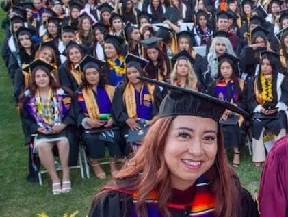 A smiling graduate in cap and gown with classmates behind her at an outdoor ceremony.