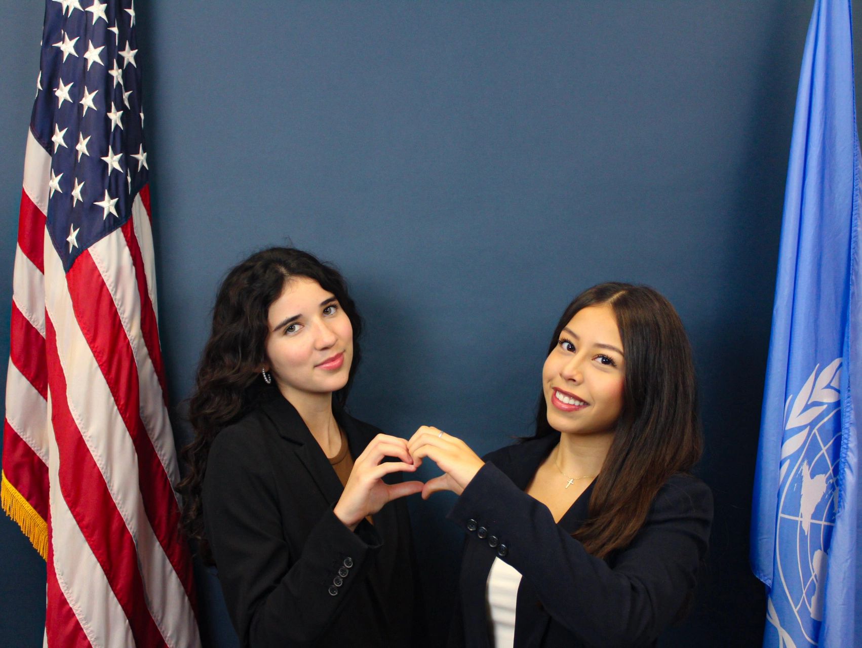 Two women forming a heart with their hands between the US and UN flags.
