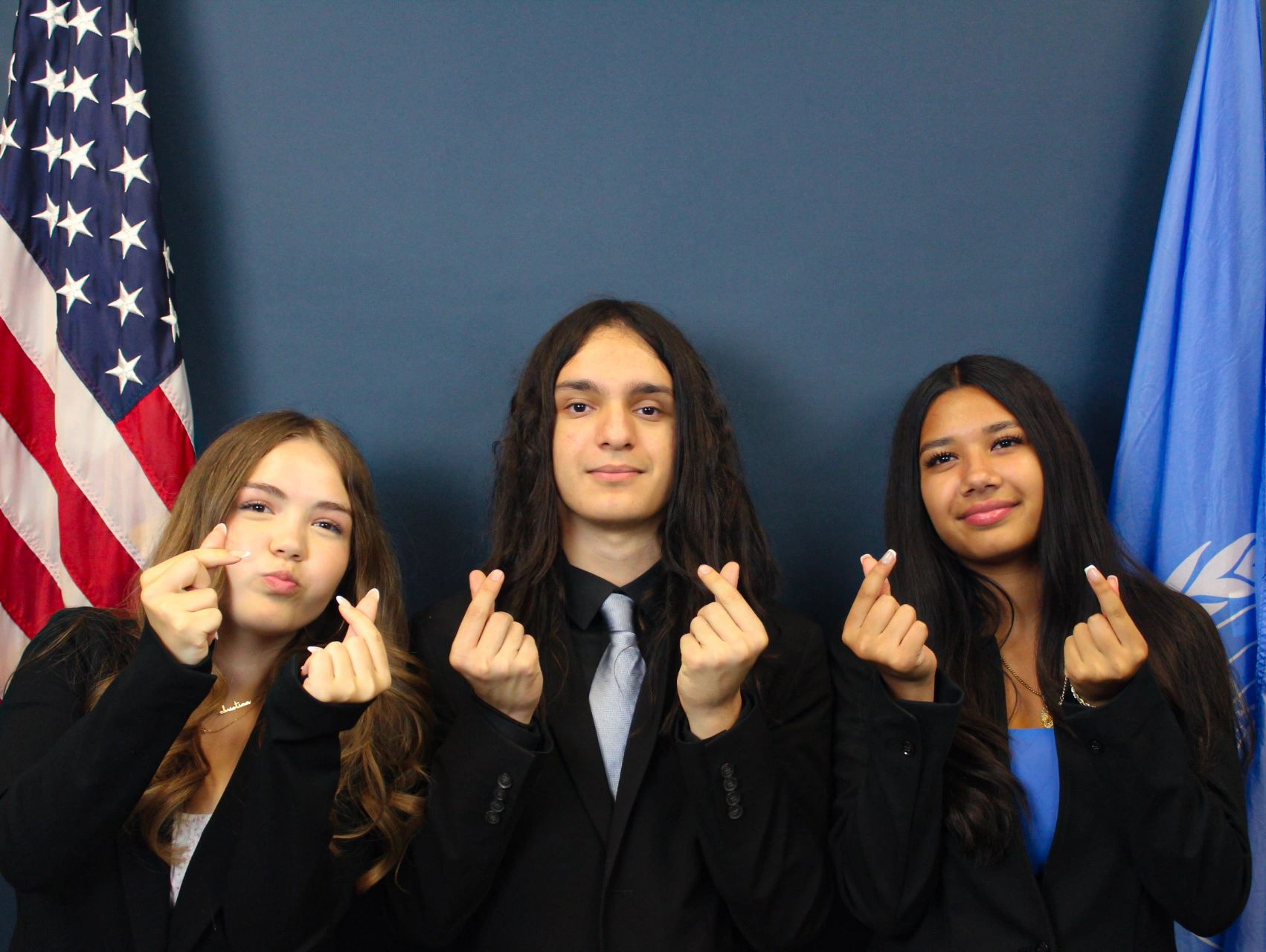Three people making finger heart gestures in front of flags.