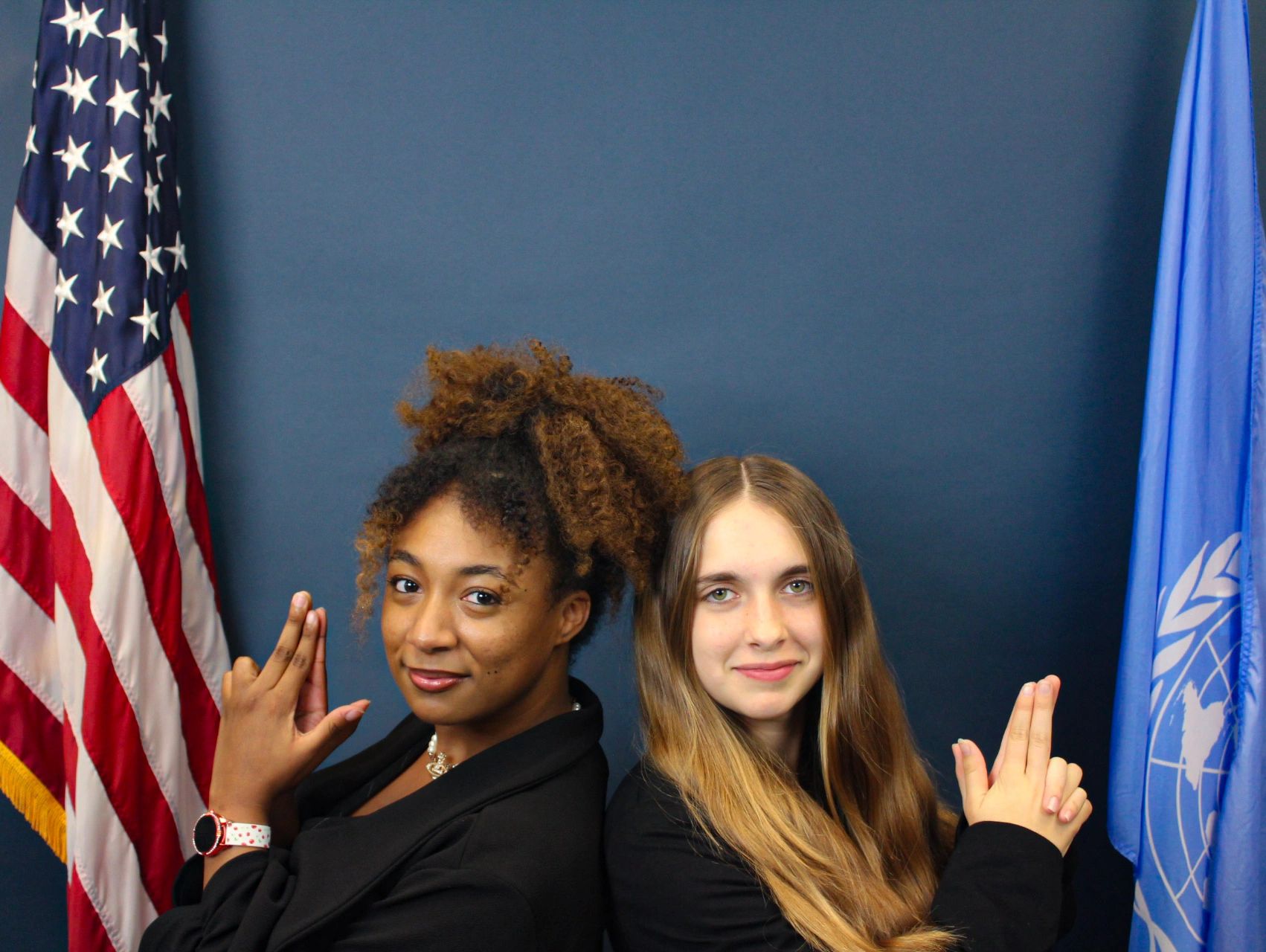 Two women posing back-to-back with playful finger guns, flags in the background.