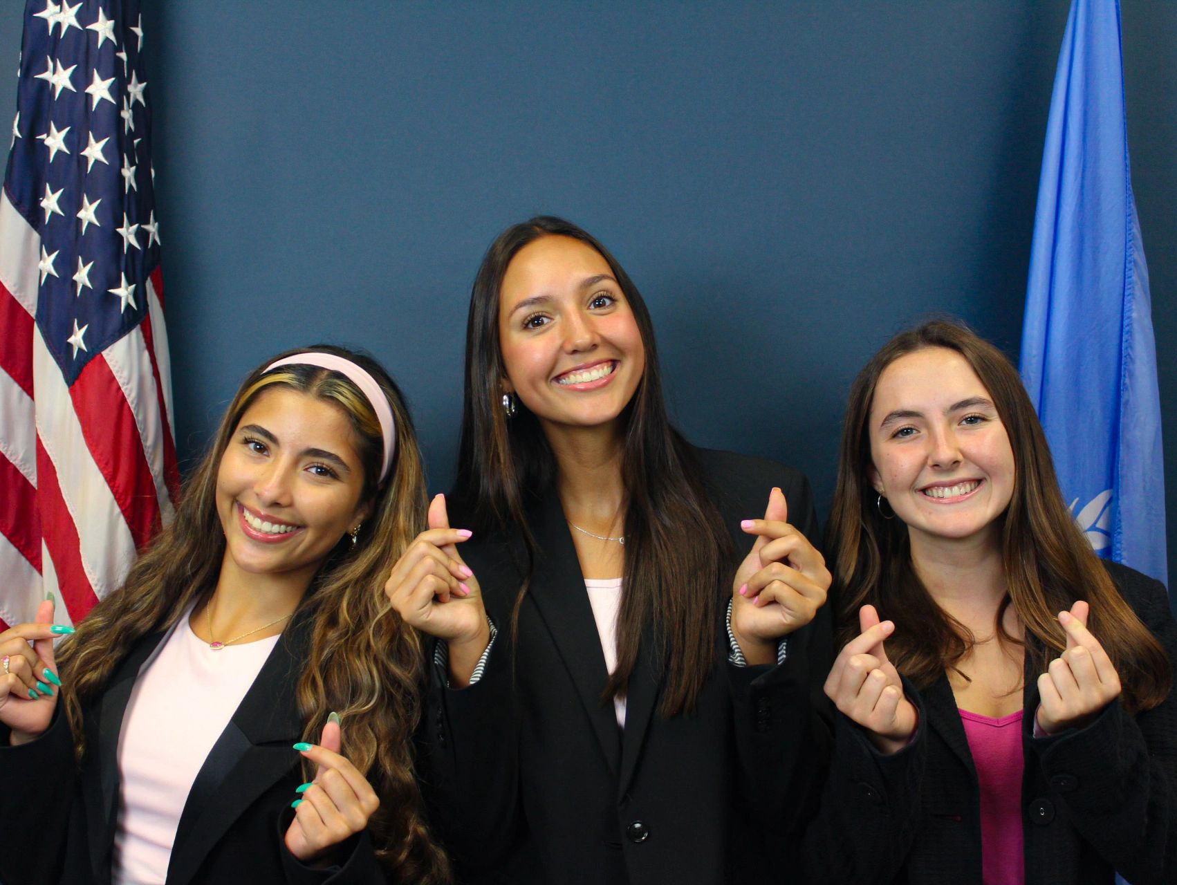 Three young women smiling and making finger heart gestures.
