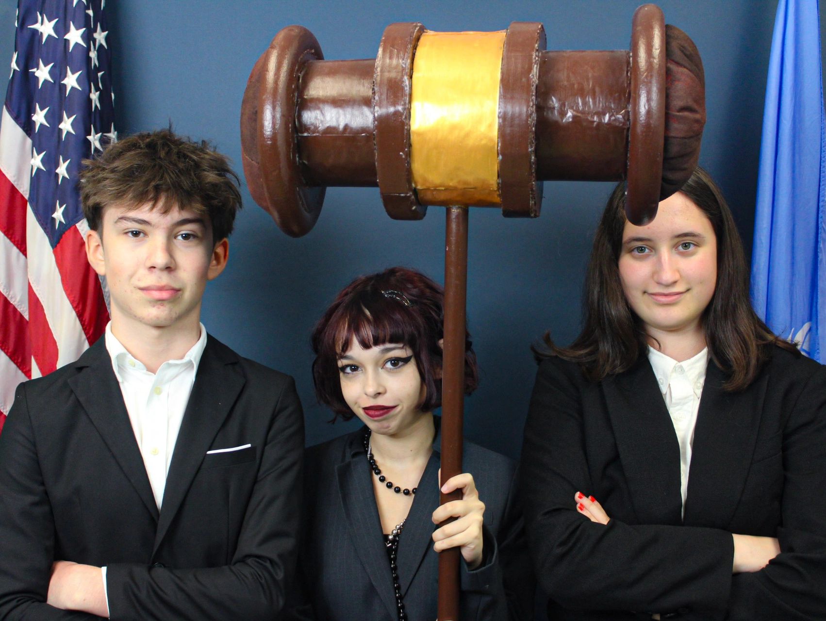 Three young people in suits with a large gavel between them.