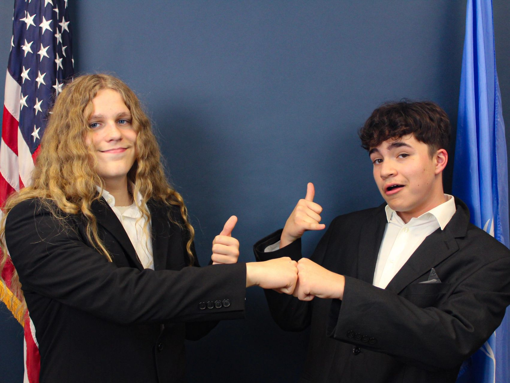 Two young men in suits fist bump with thumbs up and flags behind.