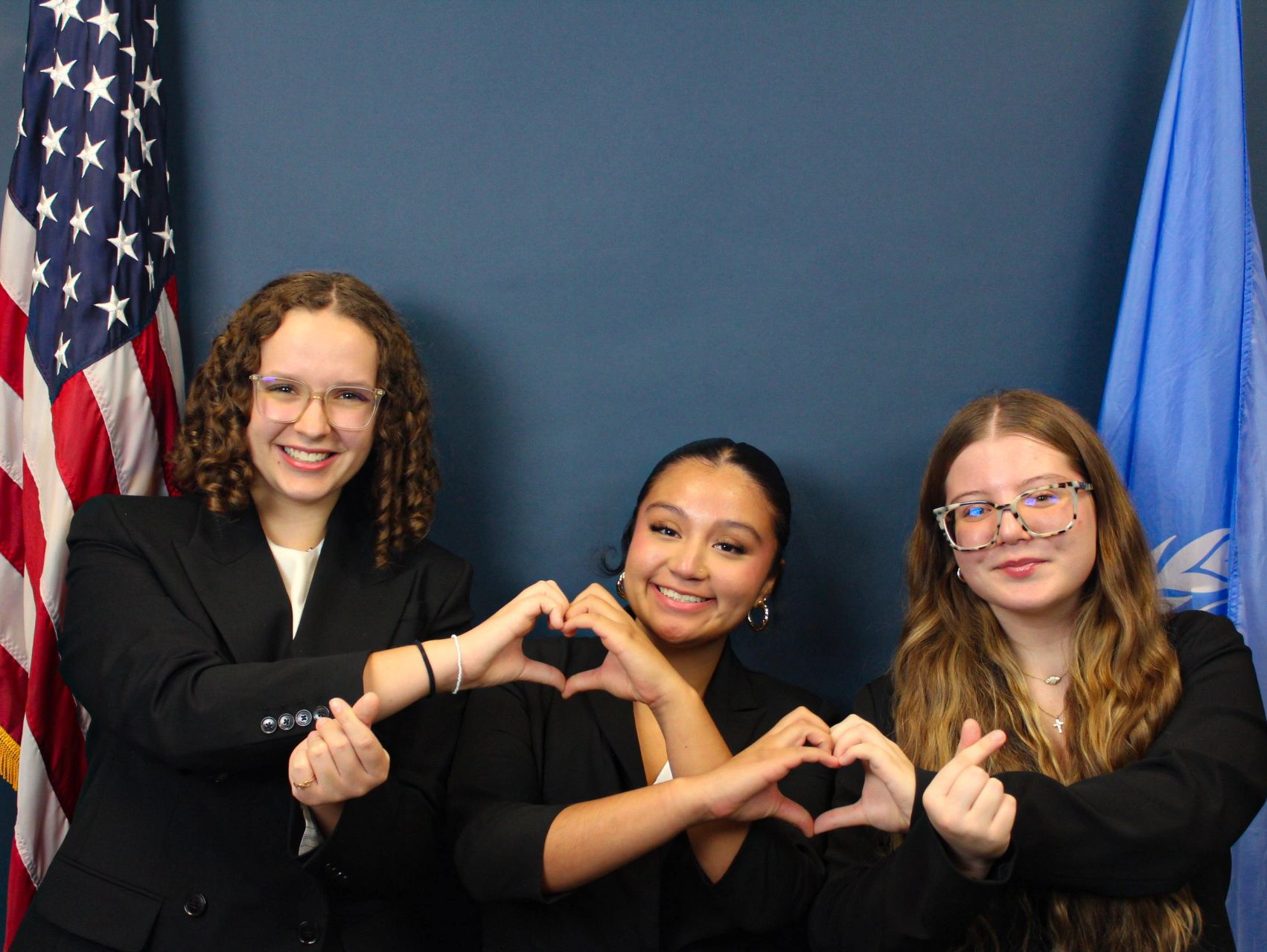 Three women making heart shapes with their hands, smiling, with flags in background.