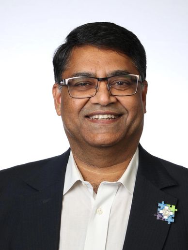 Smiling man wearing glasses and a suit jacket with a puzzle piece pin.