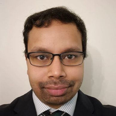 A man wearing glasses, a suit, and a striped tie smiles gently.