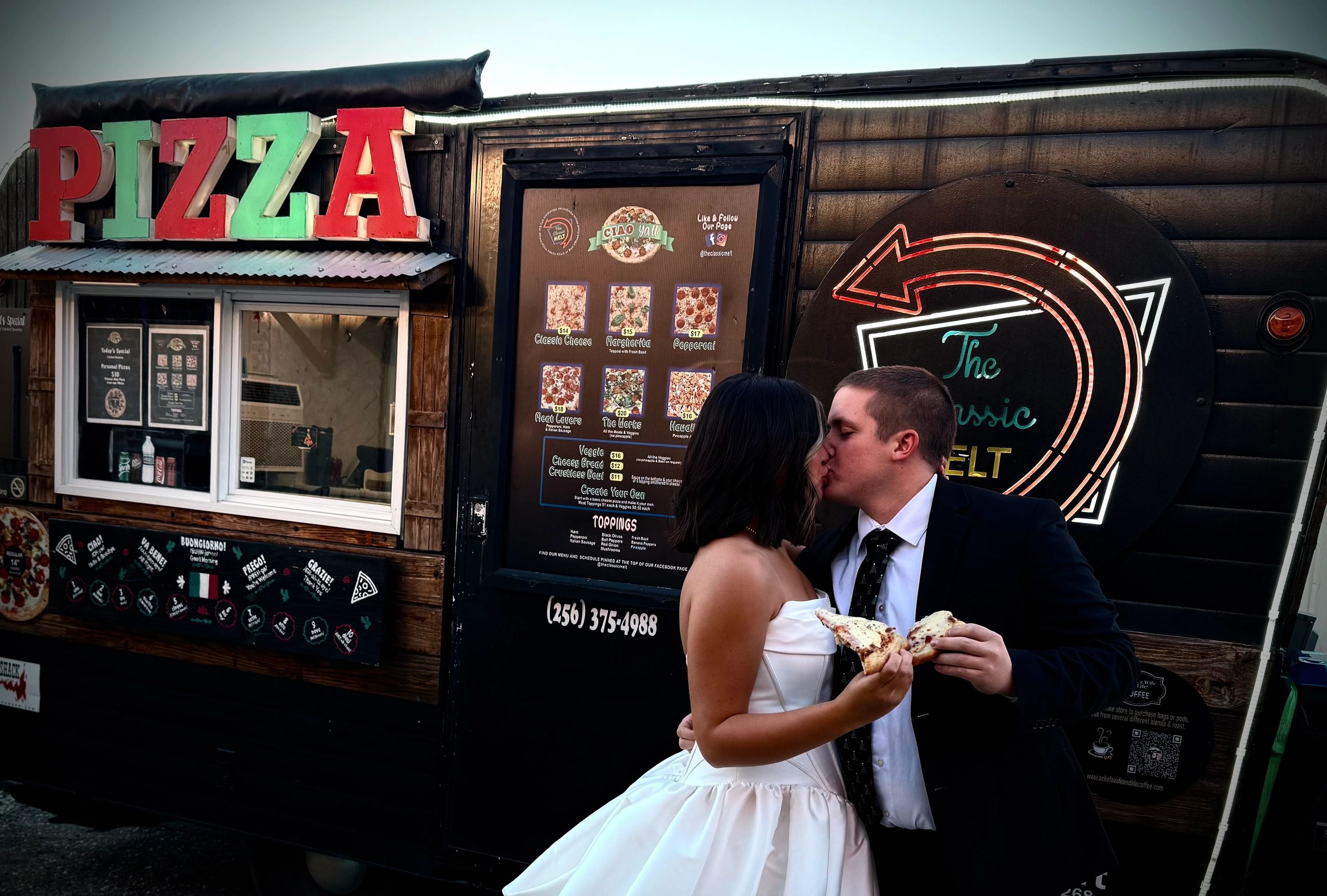 A newlywed couple shares a kiss while holding pizza slices outside a pizza food truck.