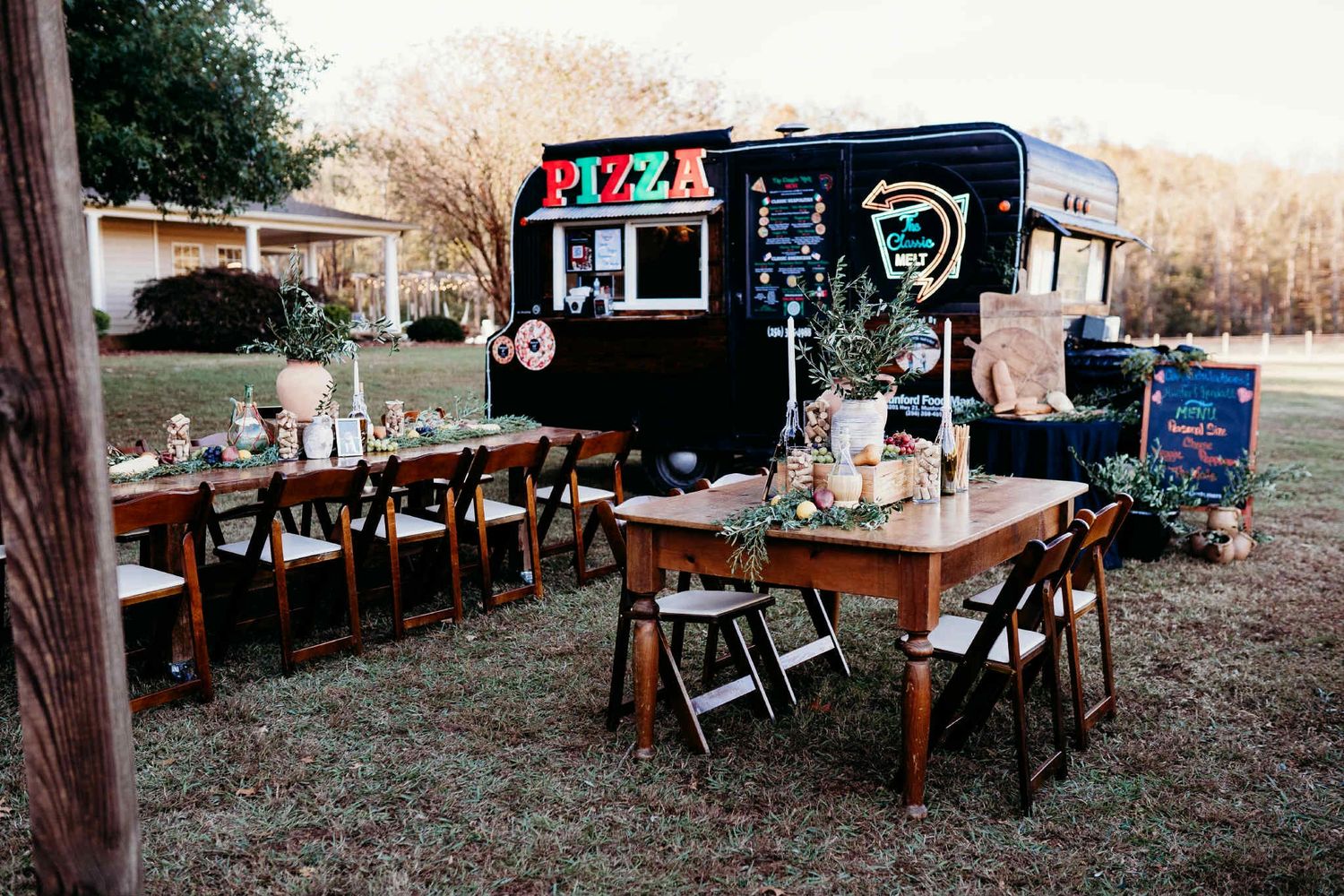 Outdoor pizza food truck with wooden tables and chairs decorated with greenery.