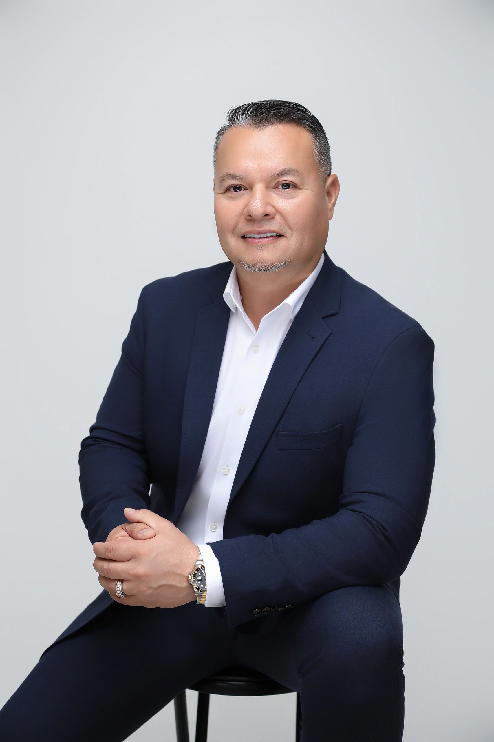 Smiling man in a navy blazer and white shirt sitting on a stool.