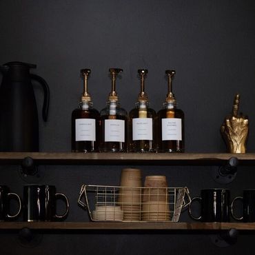 Minimalist coffee station with syrup bottles, black mugs, and disposable cups on wooden shelves.