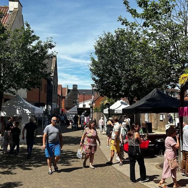 Members of the public walking down a pedestrianised street, past rows of gazebos with a blue sky.