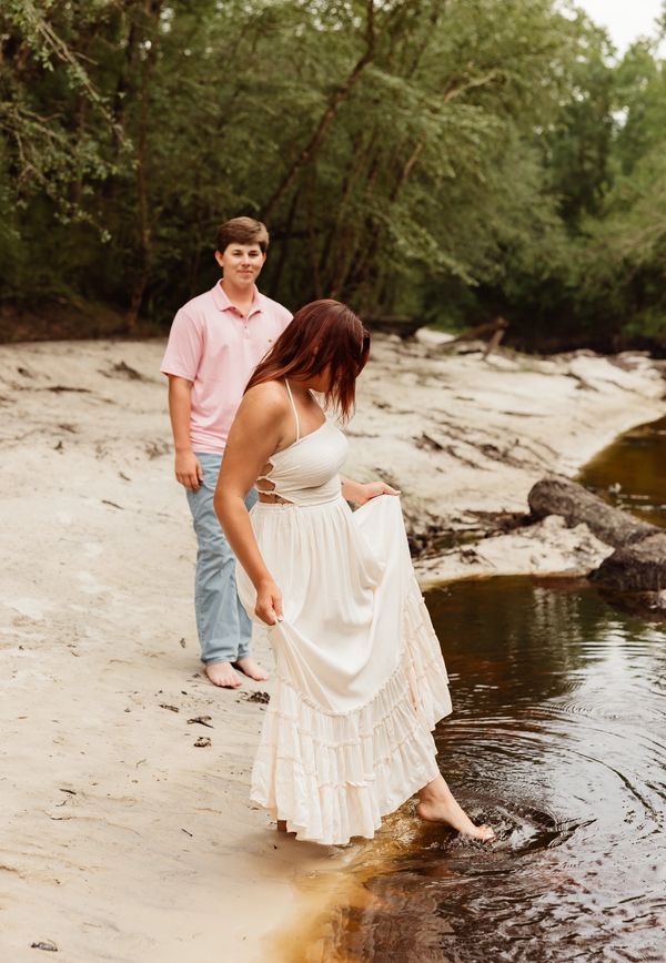 couple at river, woman is holding dress dipping toes in water, man is standing behind her smiling 