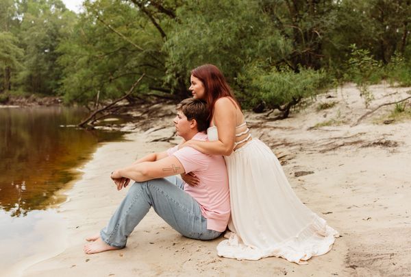 couple sitting on sandy bank of river, woman is hugging man from behind 