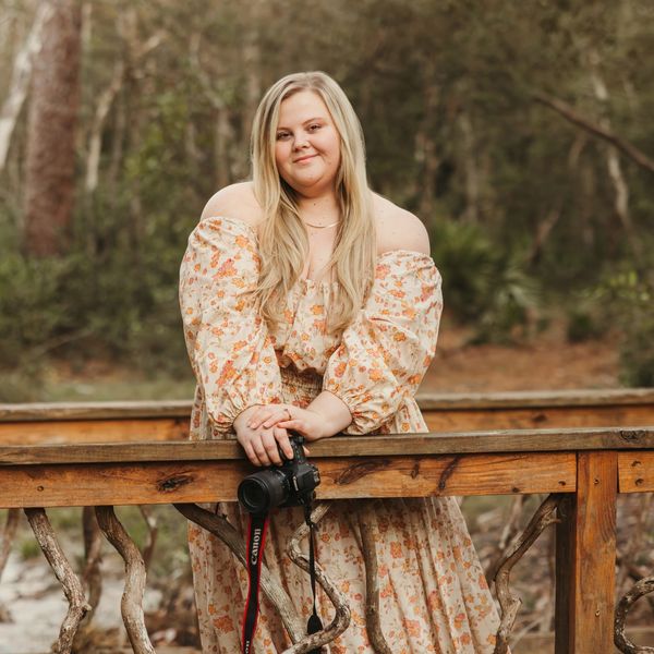 Woman in floral dress holding a camera on a wooden bridge in a forest.