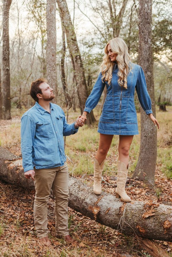 couple in forest, woman is standing on log, man is standing on ground while holding her hand smiling