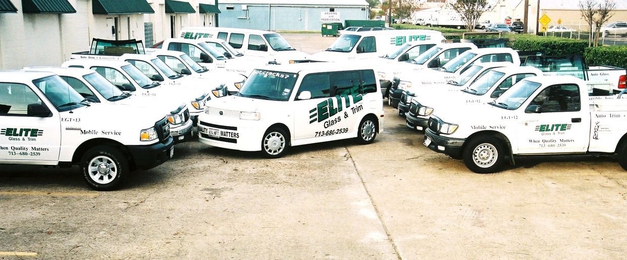 Fleet of white service vehicles branded with Elite Glass &amp; Trim logos parked outdoors.