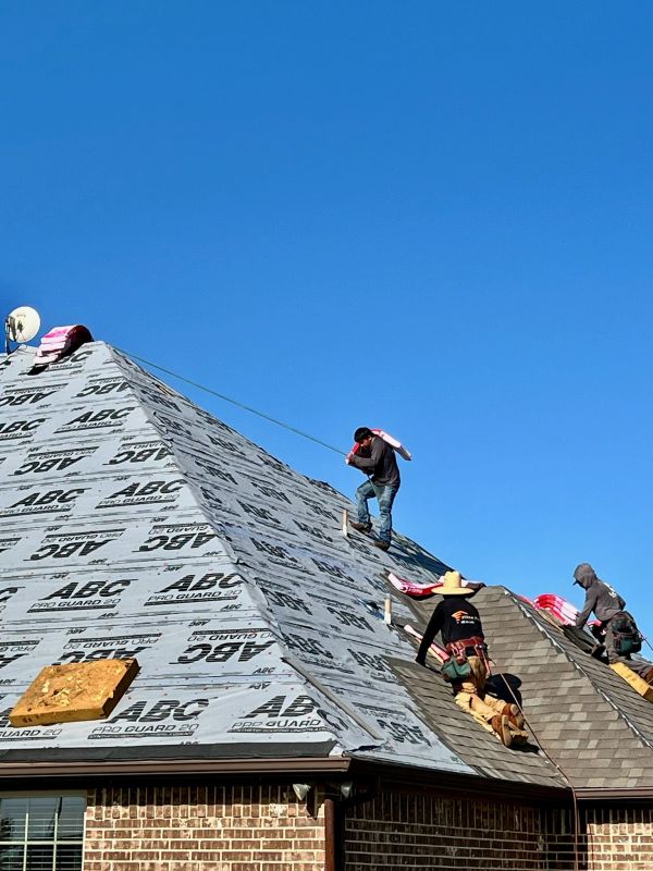 Workers installing roofing underlayment on a house roof under clear blue sky.