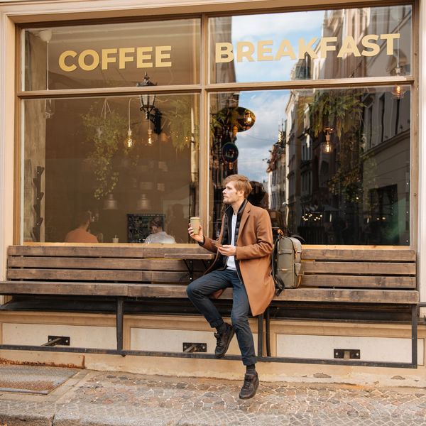 A man sitting in front of a coffee shop wearing a coat and classy clothes, and holding a coffee cup