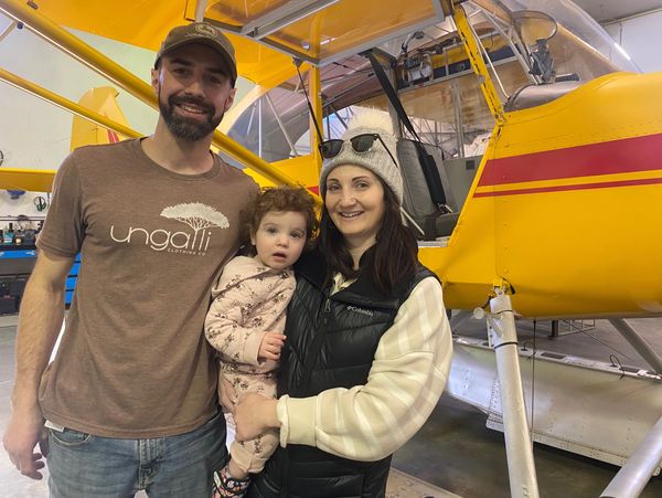 A smiling family stands in front of a yellow airplane inside a hangar.