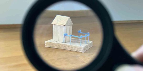 Miniatur-Holzhaus mit blauem Drahtzaun durch Lupe betrachtet.