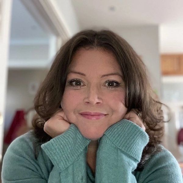 A woman smiling warmly with her hands under her chin, wearing a teal sweater.