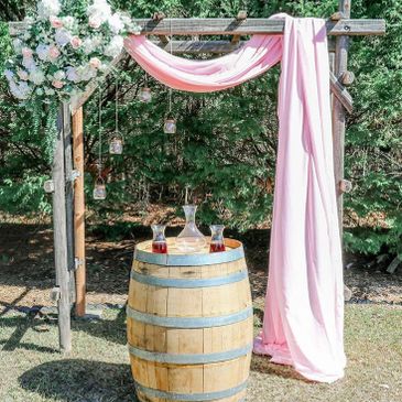 Rustic wedding arch with pink drapery and wooden barrel table outdoors.
