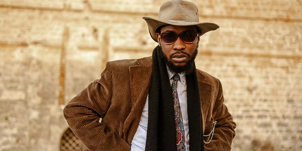 A stylish man in a brown corduroy jacket and hat poses confidently outdoors.
