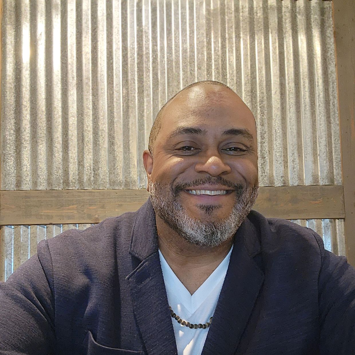 Smiling man in a navy sweater with a beaded necklace.