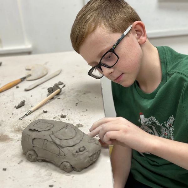 A child with black glasses looks at his work of art. He has made a car out of clay