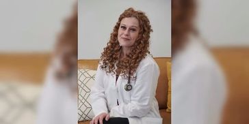A female doctor with curly hair sitting on a couch, wearing a white coat and stethoscope.