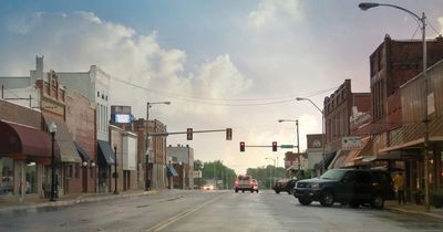 A quiet small-town street with cars and shops under a cloudy sky.