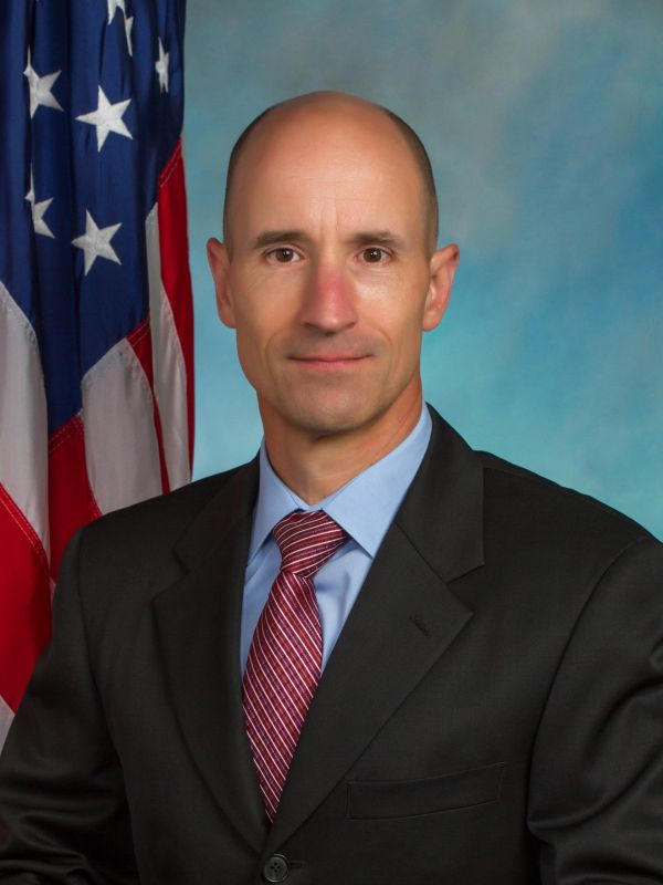 Professional portrait of a bald man in a suit with an American flag background.