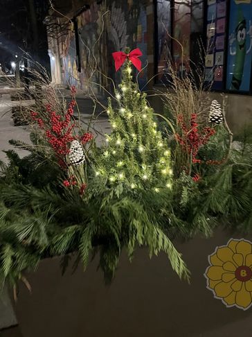 A small Christmas tree decorated with lights and a red bow, surrounded by festive greenery and pinecones.