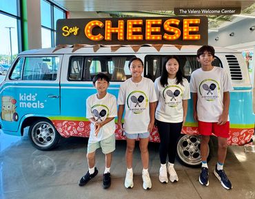 Four kids posing in front of a colorful van with a 'Say Cheese' sign.