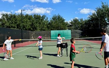 Children and a coach playing tennis on an outdoor court.