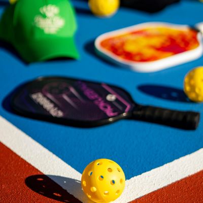 Pickleball paddle and balls on a blue court with a green cap in the background.