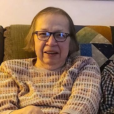 Elderly woman with glasses sitting comfortably on a couch, smiling warmly.