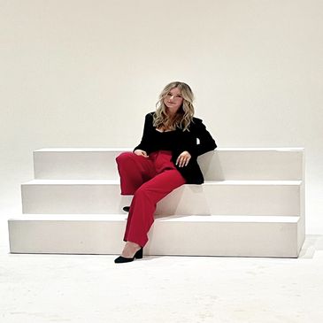 Woman in black blazer and red pants sitting on white steps.