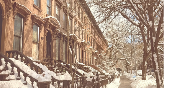 Snow-covered brownstone row houses along a clear city sidewalk in winter.